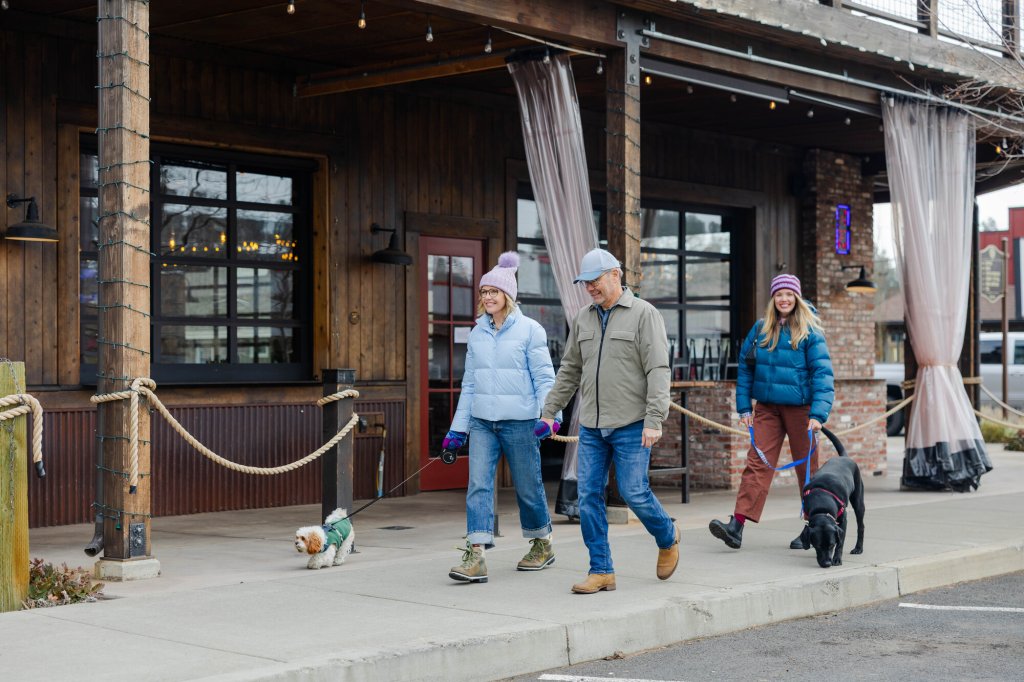 2023-01-Gritchelle-Photography-VCO-Winter-Lifestyle-Couple-190-medium (1) Three people walking two dogs on leashes along a sidewalk in front of a building in Central Oregon.