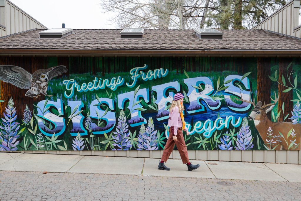 2023-01-Gritchelle-Photography-VCO-Winter-Lifestyle-Couple-166-medium A person walks past a large mural that says Greetings From Sisters Oregon in Central Oregon.