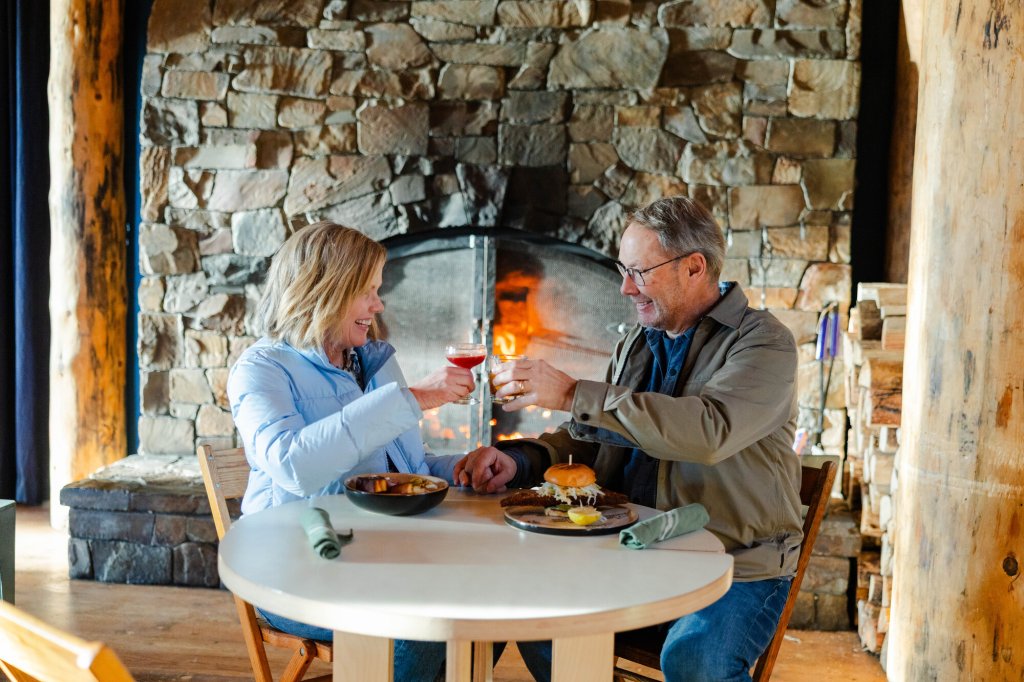 2023-01-Gritchelle-Photography-VCO-Winter-Lifestyle-Couple-145-medium (1) A couple toasts with drinks at a table in front of a stone fireplace in Central Oregon.