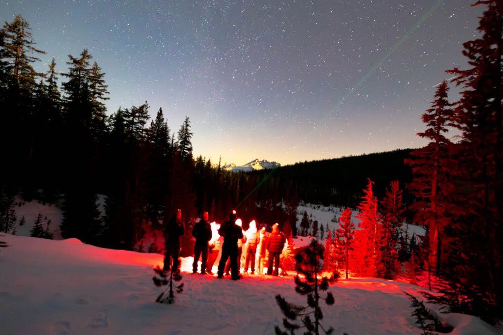 20240219-Gritchelle-Fallesgon-VCO-Bend-Winter-Lifestyle-913-medium (1) A group of people standing in snow at night under a starry sky in Central Oregon.