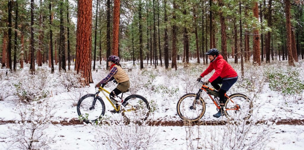 Two cyclists ride through a snowy forest, navigating a dirt path in Central Oregon.