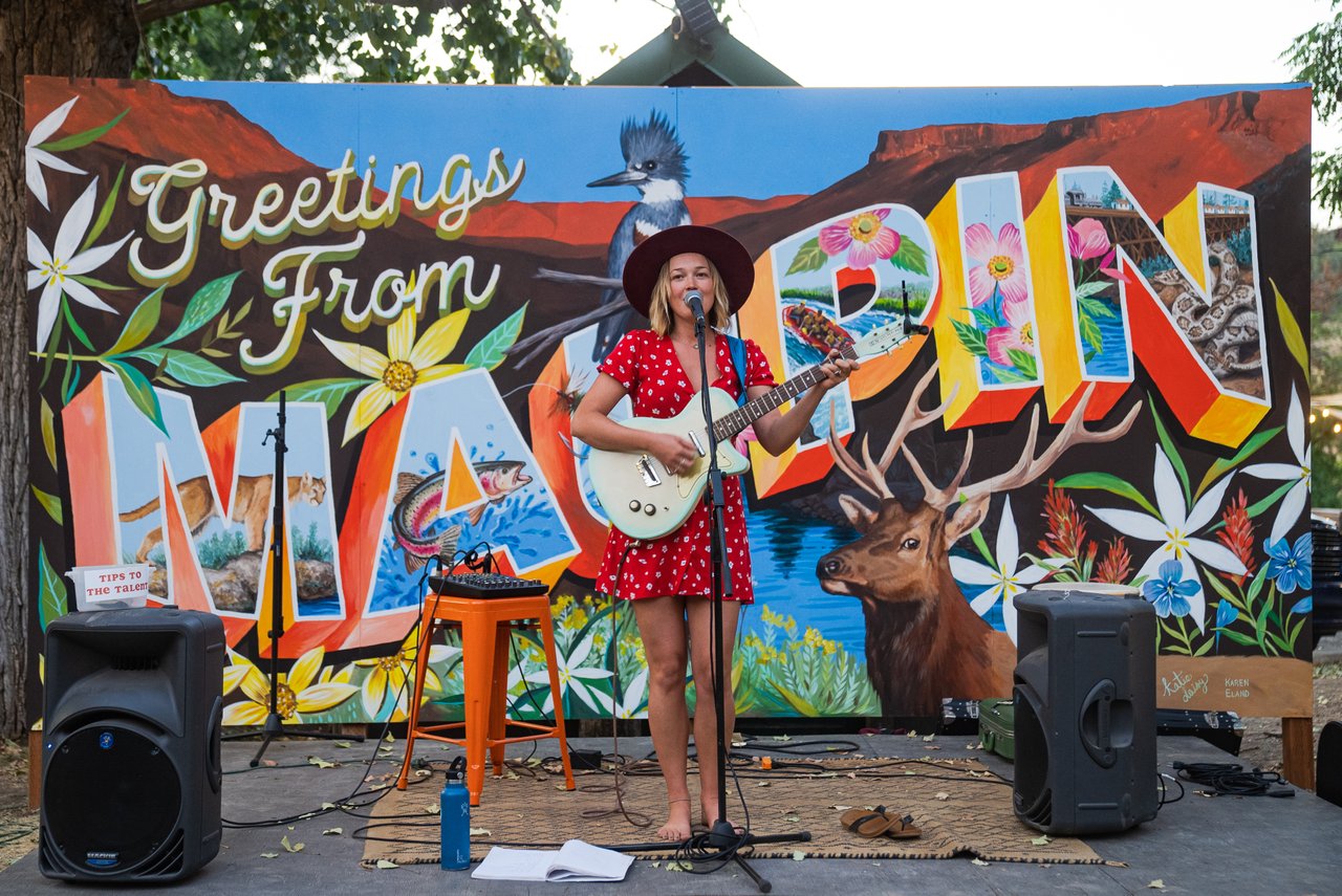 Woman in red dress and hat singing with guitar in front of a Greetings From... Mural in Maupin, oregon.