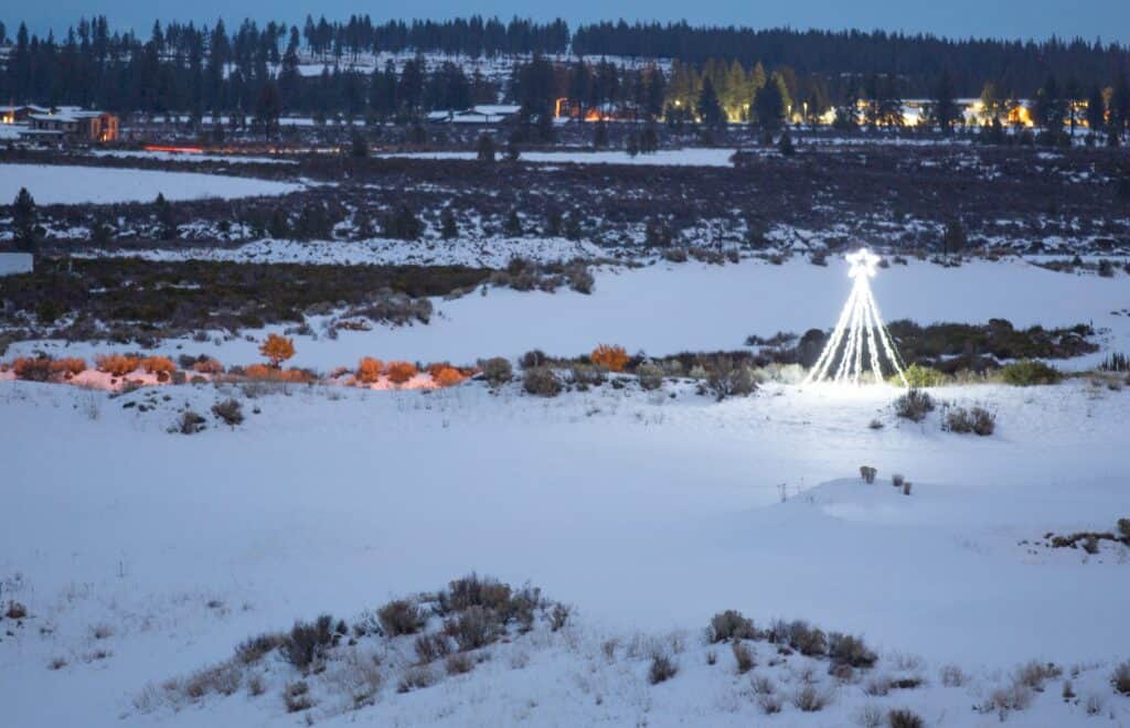 A snow-covered landscape with a Christmas tree illuminated by lights, surrounded by distant buildings and trees in Central Oregon.