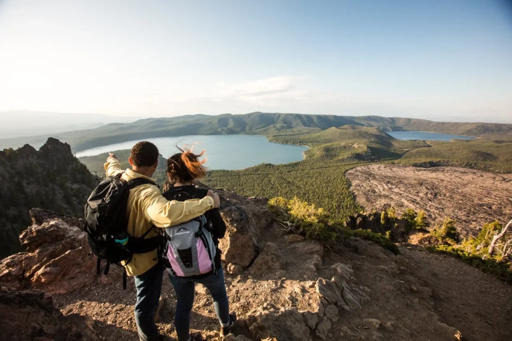 Two people stand on a trail overlooking a lake, forest, and obsidian flow.