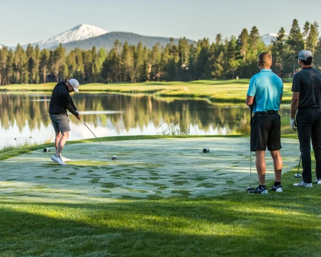 A golfer hits a golf ball with a pond, pine forest, and mountain in the background.