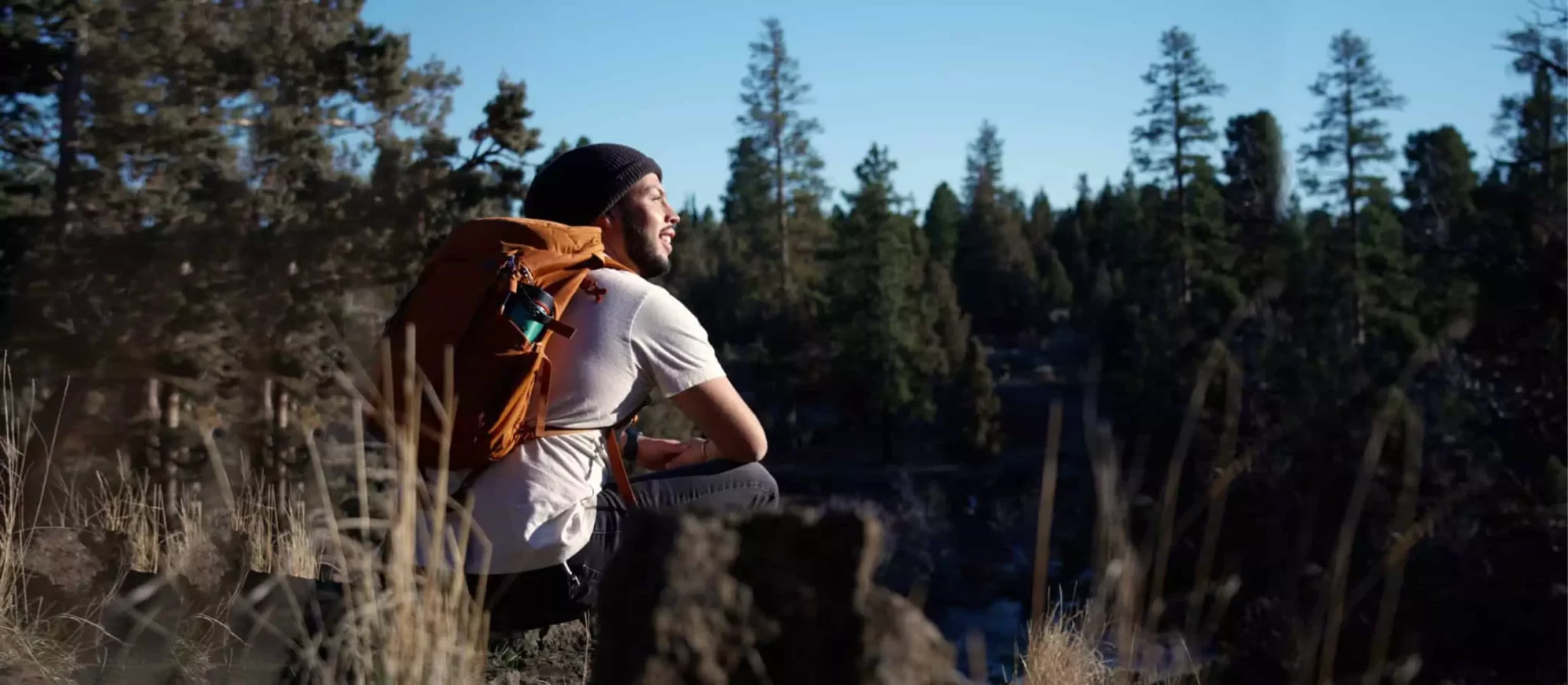 A person with a backpack sits on a rocky outcrop, surrounded by tall trees and wildflowers, gazing at the clear blue sky in Central Oregon.