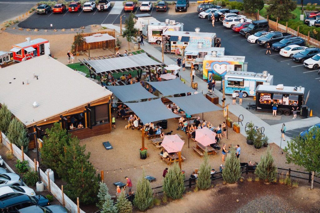 An overhead view of several people seated at an outdoor food truck park and several food trucks and cars parked on the adjacent street.