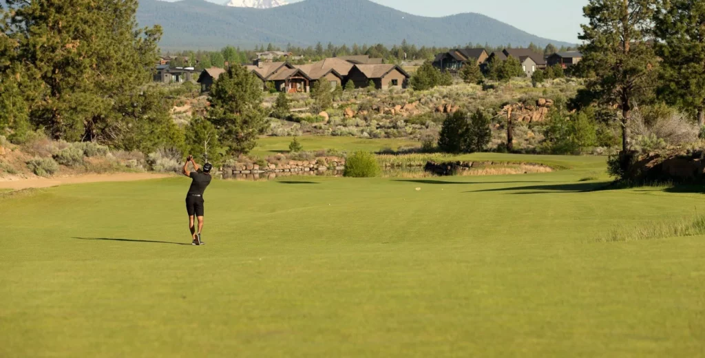 A golfer in mid-swing on a lush green golf course with houses and mountains in the background in Central Oregon.