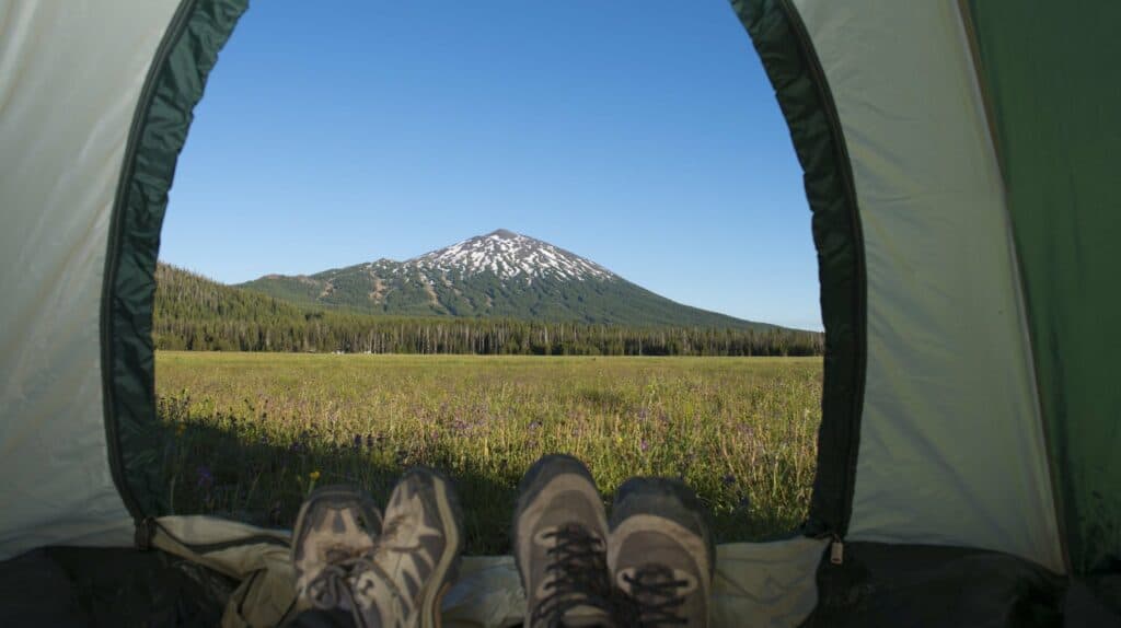 A person's feet peek out of a tent, overlooking a vast meadow and a snow-capped mountain in Central Oregon.
