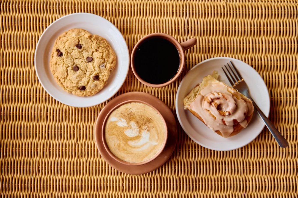 A cozy breakfast spread featuring a chocolate chip cookie, coffee, cinnamon roll, and cappuccino, all arranged on a woven table mat in Central Oregon.