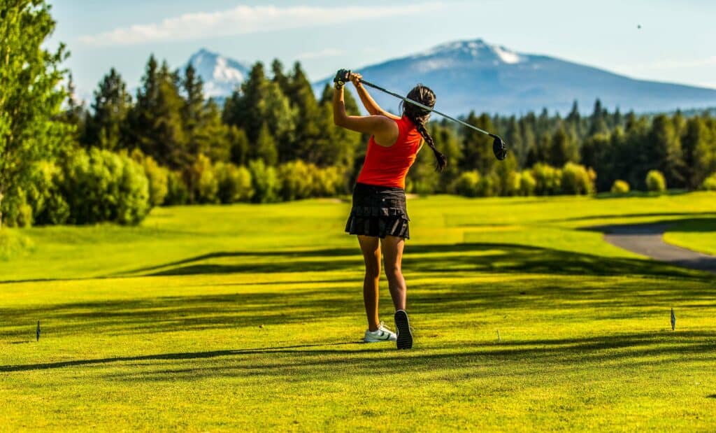 A woman in a red top and black skirt plays golf on a green field with mountains in the background in Central Oregon.