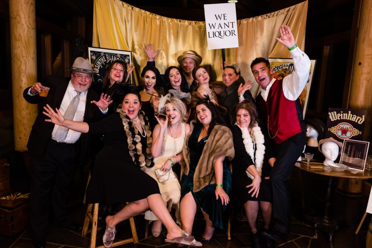 A lively group posing for a photo at a 1920s-themed party, with a 'We Want Liquor' sign and vintage decor in Central Oregon.