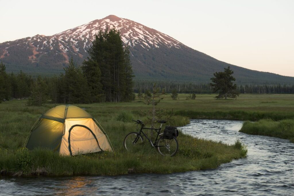 A serene camping scene at dusk with a tent, bicycle, and flowing river beside a snow-capped mountain in Central Oregon.