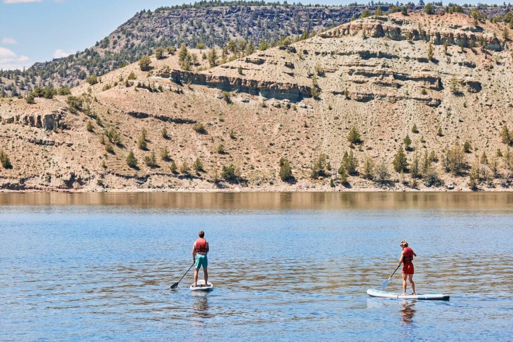 Two people paddleboarding on a calm lake surrounded by desert hills in Central Oregon.