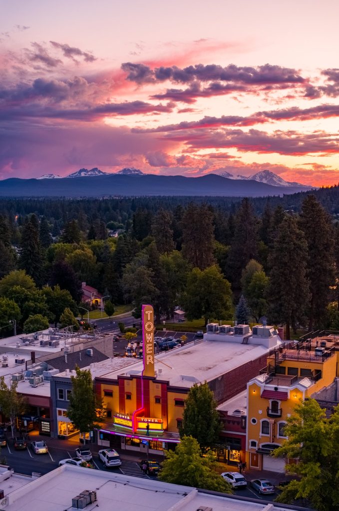 A vibrant cityscape at sunset, featuring a historic theater with a neon sign, surrounded by lush greenery and distant mountains in Central Oregon.