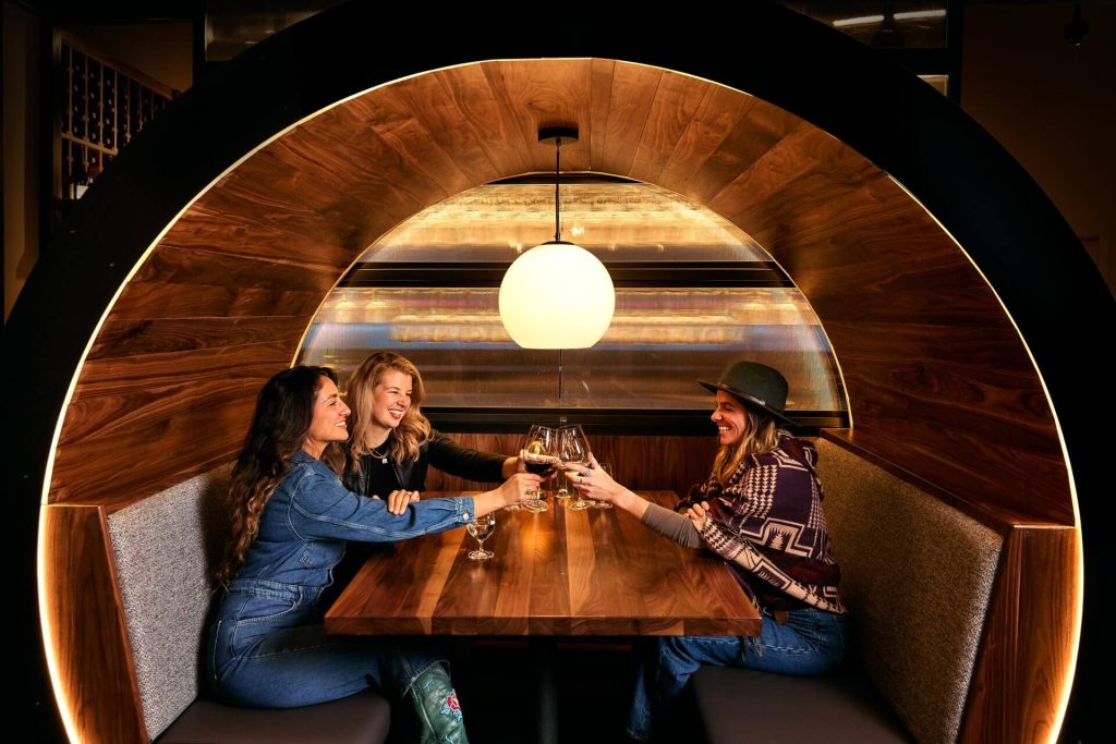 Three friends enjoying a toast in a cozy, circular booth with warm lighting in Central Oregon.