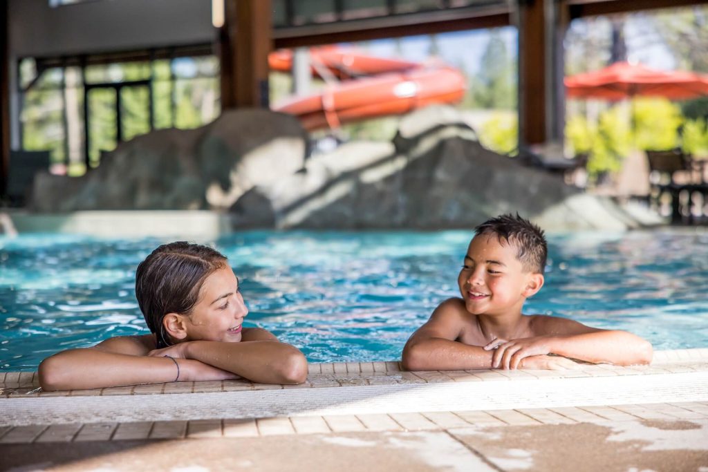 Two children enjoying a swim in an indoor pool, surrounded by lush greenery and a colorful slide in Central Oregon.