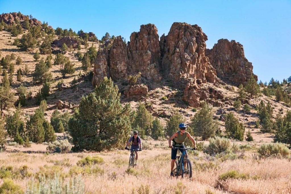Two mountain bikers ride through a dry, grassy landscape with rocky outcrops in Central Oregon.