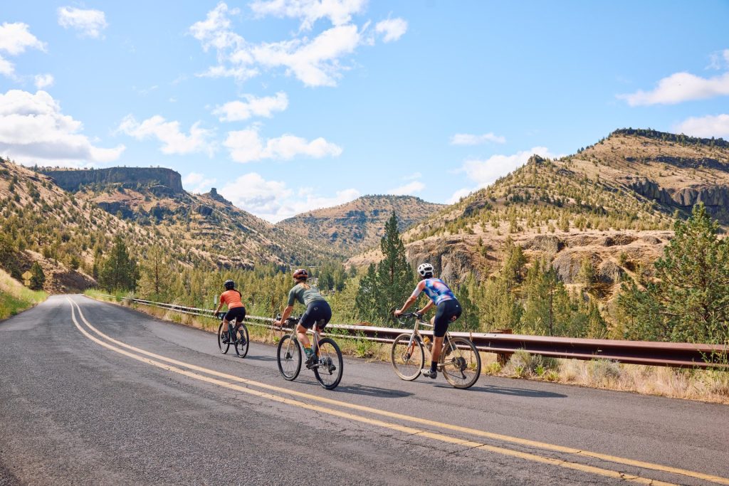 Three cyclists ride down a winding road surrounded by mountains and trees in Central Oregon.