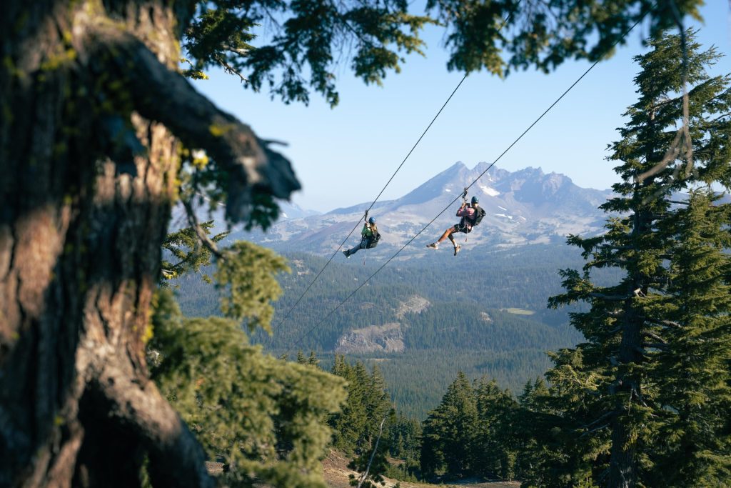 two people enjoy separate, parallel ziplines as they gaze at the mountains in Central Oregon
