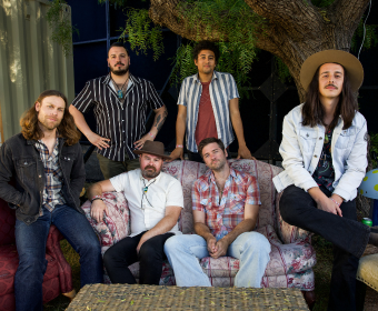 A group of seven people, including three men standing and four seated, pose together in a relaxed outdoor setting in Central Oregon.