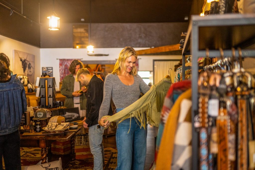 A woman in a grey long-sleeved shirt and blue jeans holds up and examines a pea-green jacket in a clothing boutique. The jacket is among other western-themed clothing items for sale.