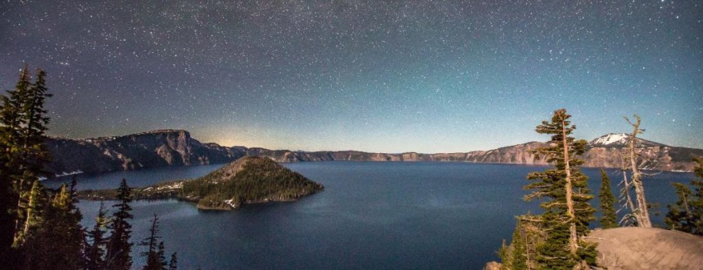 A serene night sky over a starry lake with mountains and trees in Central Oregon.