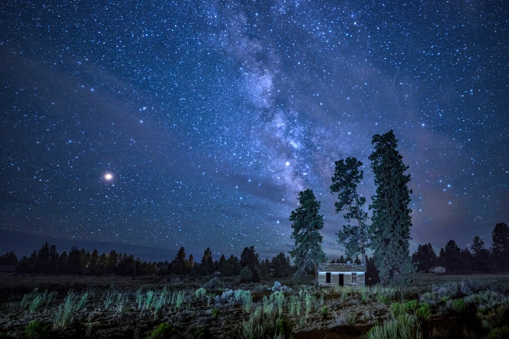 A serene night scene featuring a small wooden cabin surrounded by tall trees under a starry sky in Central Oregon.