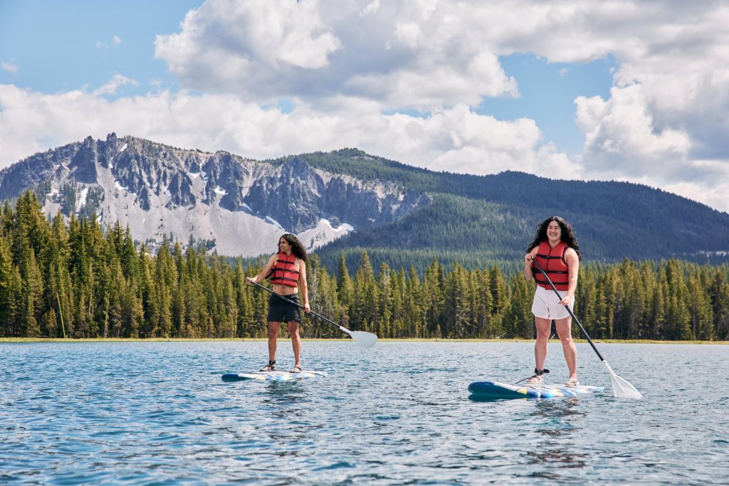 Two women paddleboarding on a serene lake surrounded by towering mountains and lush forests in Central Oregon.