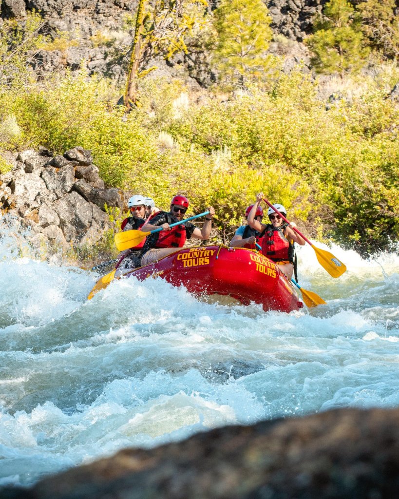 Four adventurers in a red raft navigate a whitewater river, wearing helmets and life jackets in Central Oregon.