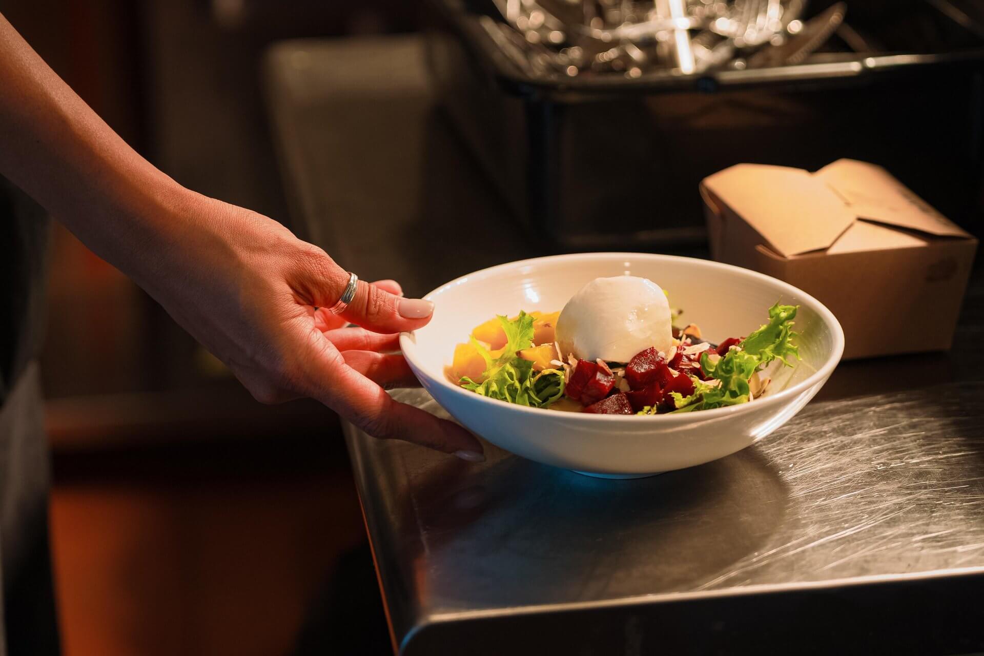 A hand holding a bowl of salad with beets and a dollop of cream, placed on a stainless steel counter in Central Oregon.