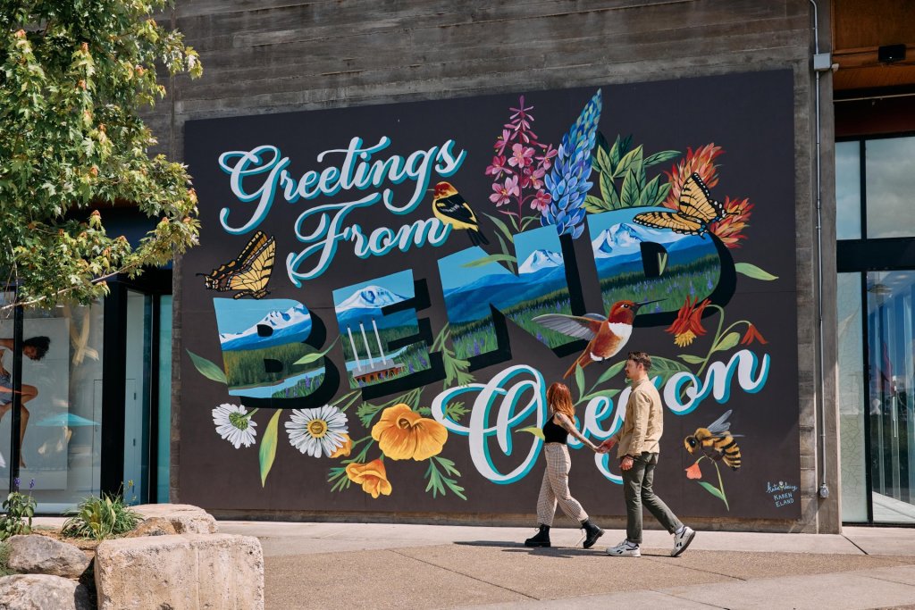 Two people walk past a vibrant mural on a building wall, featuring nature and the text 'Greetings from Bend, Oregon' in Central Oregon.