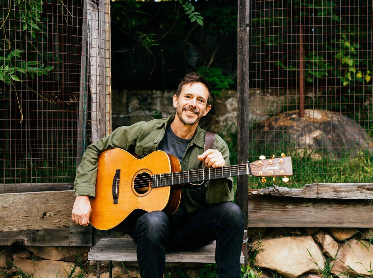 A man with a guitar, smiling, sitting on a rustic bench in Central Oregon.