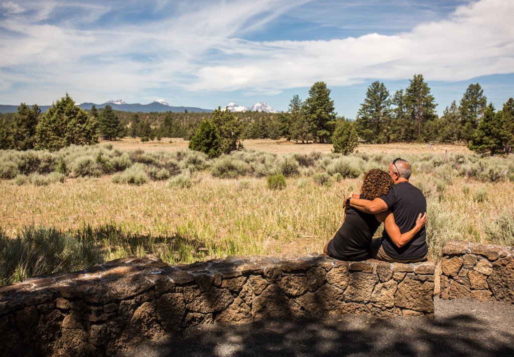 A couple sits on a stone wall, gazing at a vast, grassy landscape with distant mountains in Central Oregon.