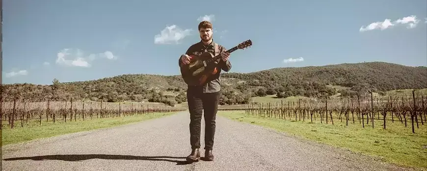 A man walks down a winding road with a guitar, surrounded by vineyards and rolling hills in Central Oregon.