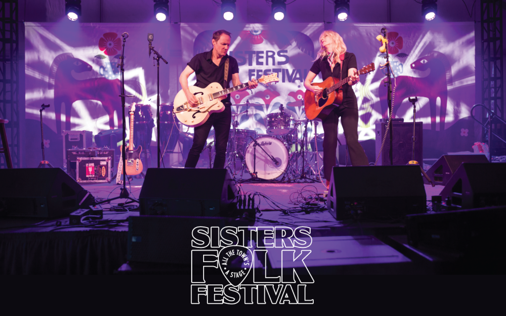 Two musicians perform on stage at the Sisters Folk Festival, playing guitars and drums under vibrant purple lights in Central Oregon.
