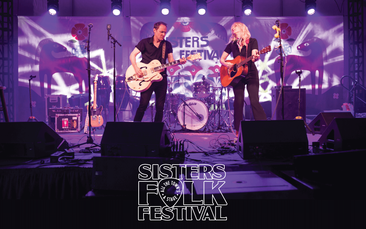 Two musicians perform on stage at the Sisters Folk Festival, playing guitars and drums under vibrant purple lights in Central Oregon.