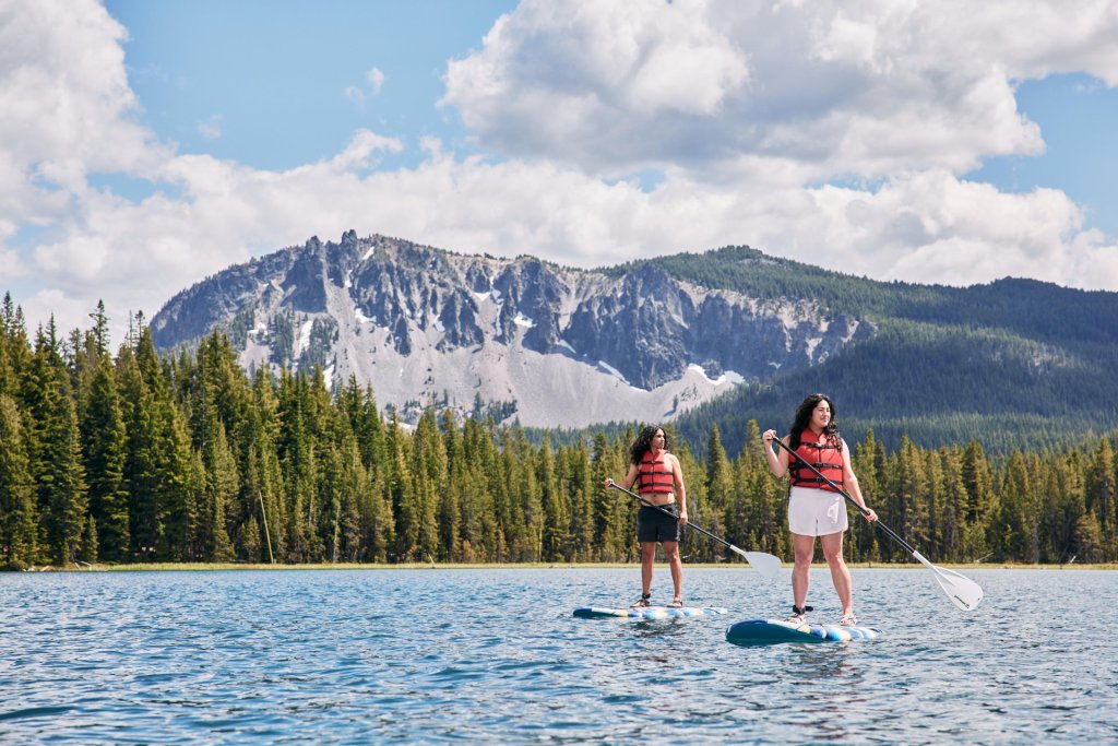 Two women paddleboarding on a serene lake surrounded by towering mountains and lush forests in Central Oregon.