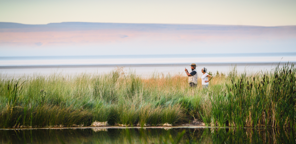 Two people standing in tall grass near a serene lake, gazing at distant mountains in Central Oregon.