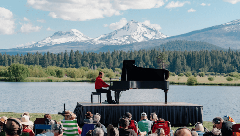 A musician plays a grand piano on a stage overlooking a serene lake surrounded by mountains and trees in Central Oregon.