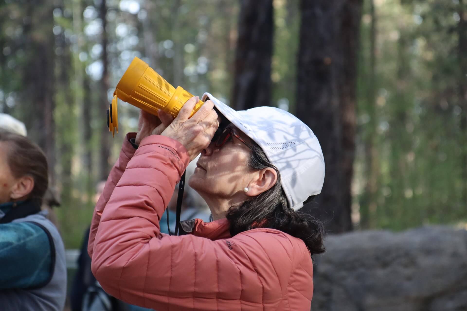 A woman in a red jacket and white hat uses a yellow telescope in a forest in Central Oregon.