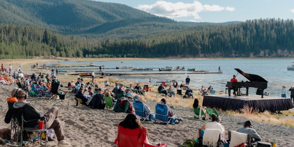 A lively outdoor concert by a grand piano on a lakefront stage, surrounded by a crowd enjoying the music and scenic views in Central Oregon.