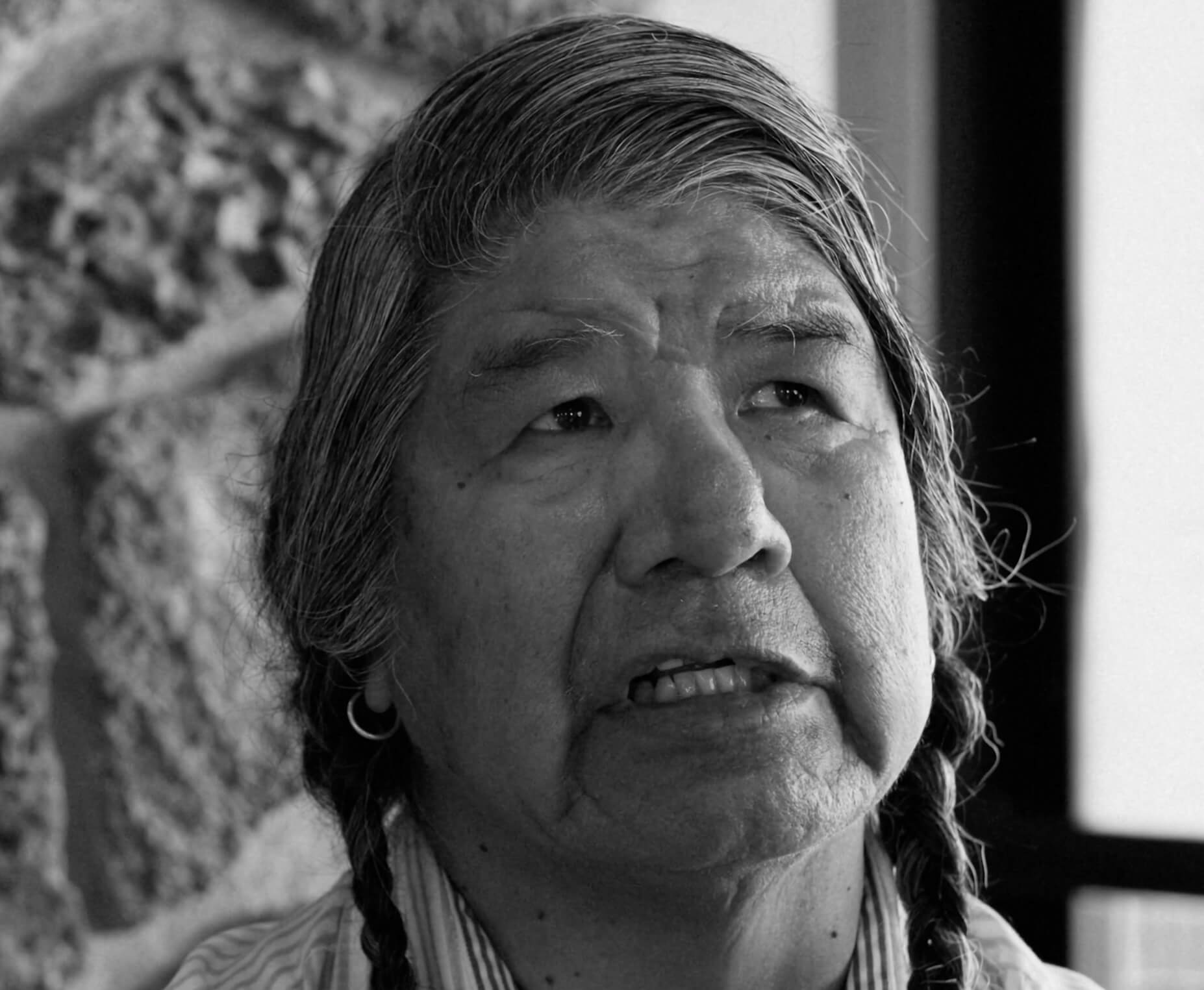 An elderly woman with long braids and a thoughtful expression, standing in front of a stone wall in Central Oregon.