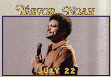 Trevor Noah holding a microphone, smiling, dressed in a brown jacket, ready to perform in Central Oregon.