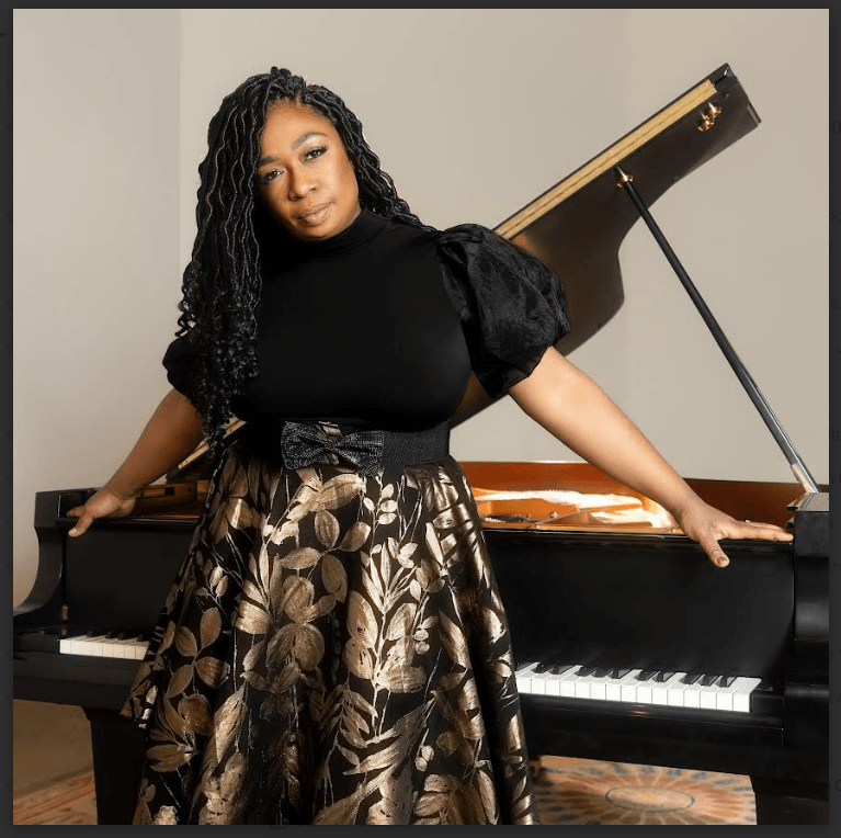 A woman with long, braided hair sits elegantly at a grand piano, dressed in a stylish black top and a gold floral skirt in Central Oregon.