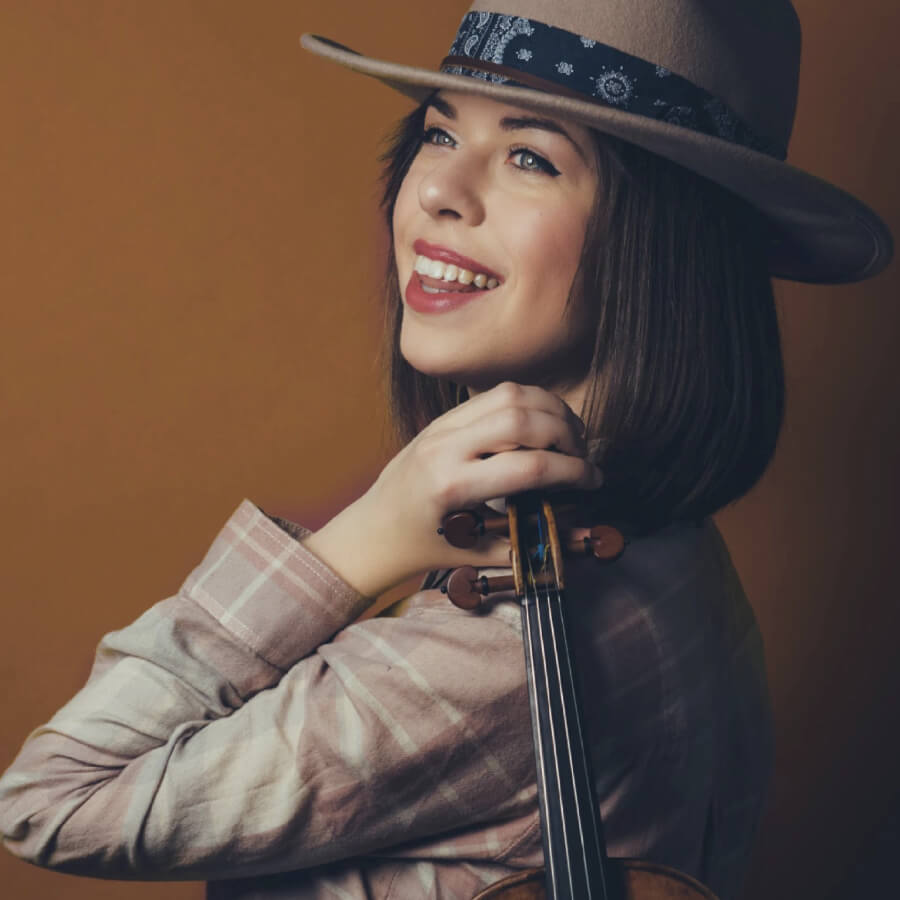 A woman with a hat and violin, smiling warmly in Central Oregon.