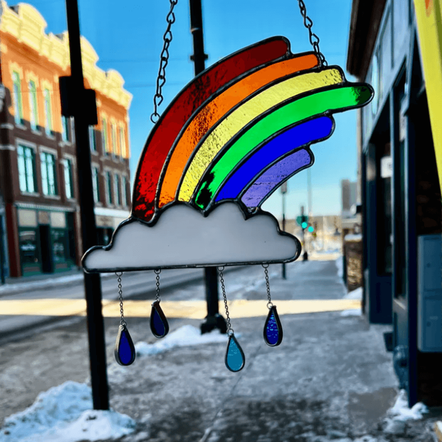 A vibrant rainbow-colored stained glass cloud with teardrop rain, hanging on a snowy street in Central Oregon.
