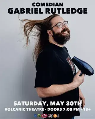 A bearded man with glasses and a black shirt holds a hairdryer, his long hair blowing in the wind in Central Oregon.