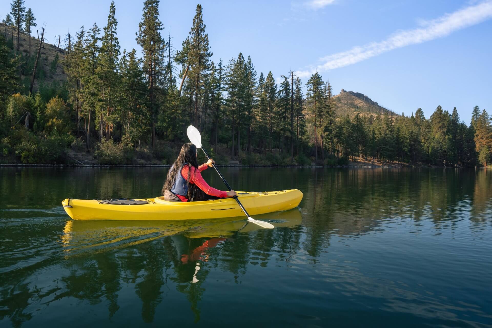 A woman paddles a yellow kayak on a serene lake surrounded by tall trees in Central Oregon.