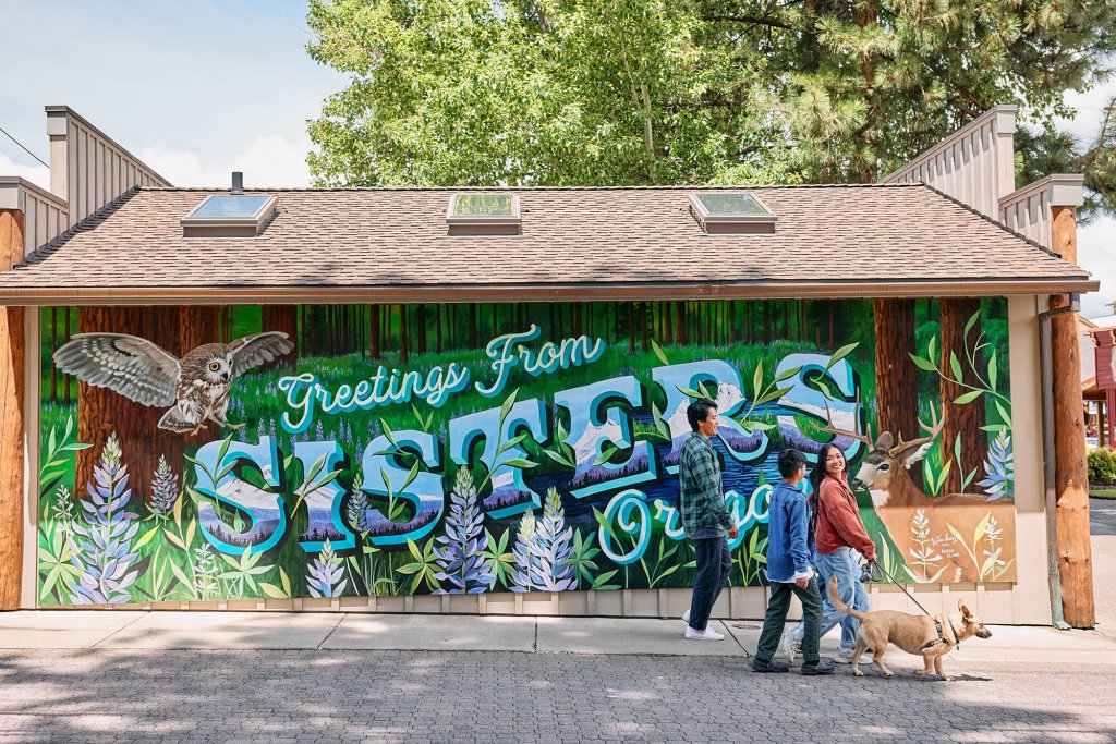 People and a dog walk past a vibrant mural featuring nature and the word 'Sisters' in Oregon in Central Oregon.
